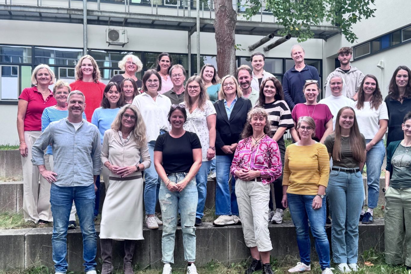 Gruppenfoto von 28 Personen im Freien, lächelnd und stehend auf einer Treppe.