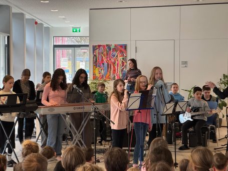 Schülergruppe führt ein Musikstück in einem Raum mit buntem Kunstwerk auf.