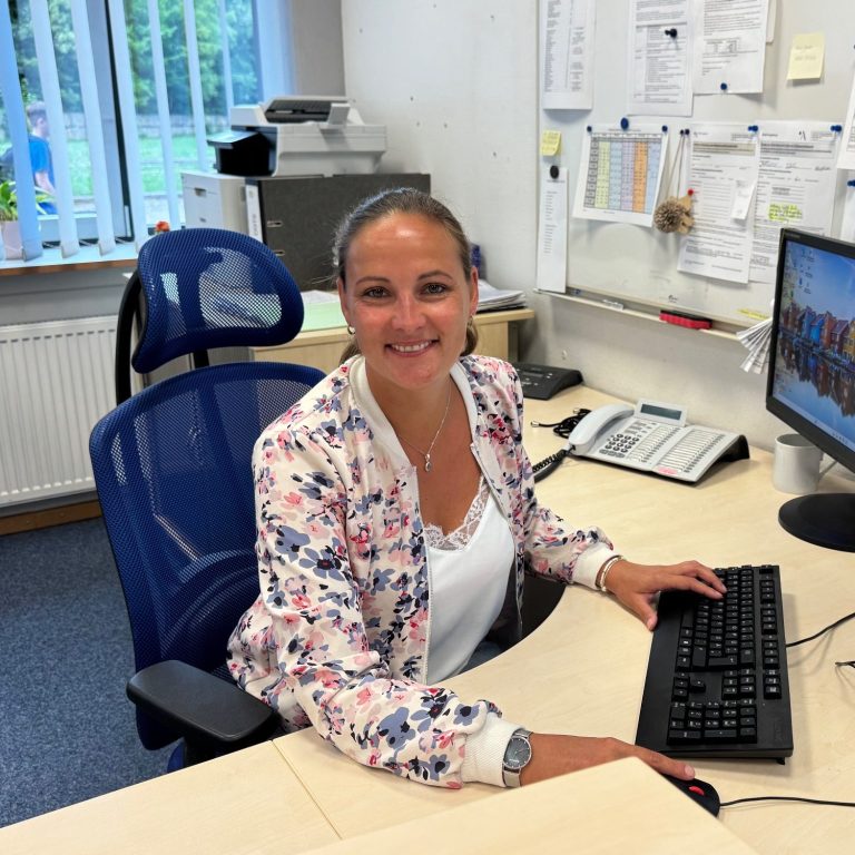Frau lächelt am Schreibtisch vor einem Computer in einem Büroumfeld.