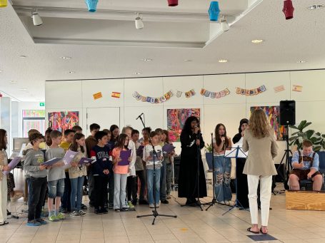 Schüler singen in einem festlichen Raum, begleitet von Lehrern und Instrumenten.