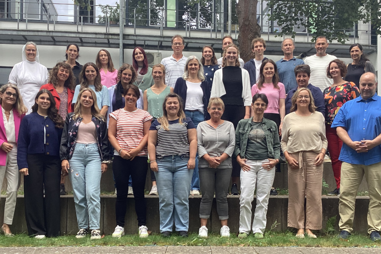 Gruppenfoto von 28 Personen im Freien, lächelnd und stehend auf einer Treppe.