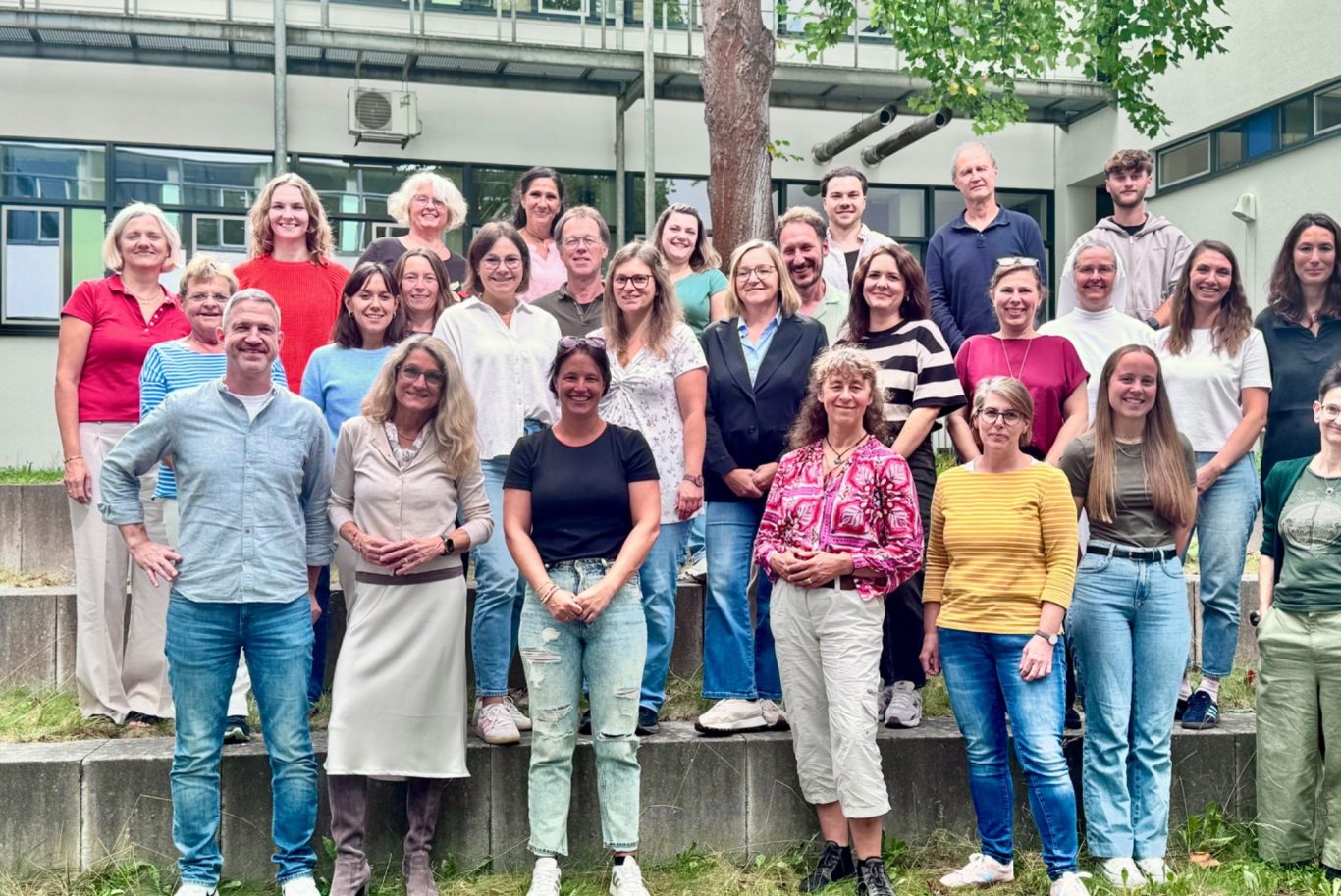 Gruppenfoto von 28 Personen im Freien, lächelnd und stehend auf einer Treppe.
