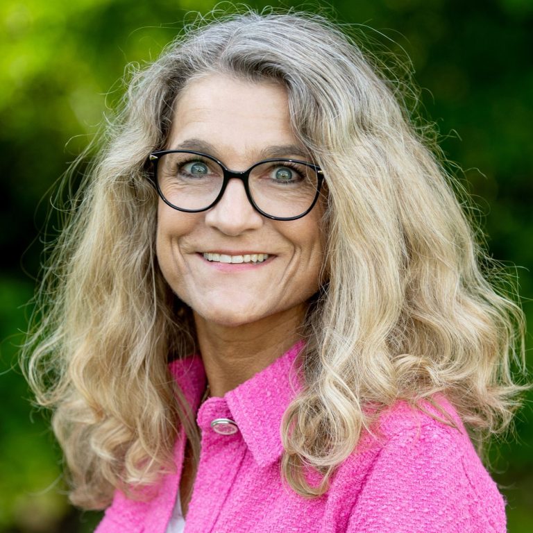 Frau mit langen, lockigen Haaren und Brille, lächelt vor grünem Hintergrund.